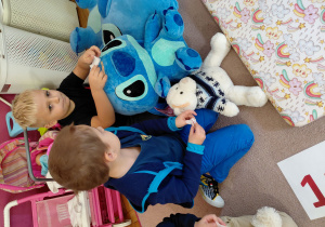 Chłopcy siedząc przyklejają plastry pluszakom.