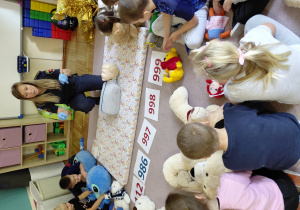 Przedszkolaki siedzą z pluszakami przed kartonikami z numerami alarmowymi. Na dywaniku siedzi strażniczka miejska. Przed nią leży manekin do ćwiczeń resuscytacji.