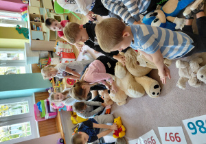 Przedszkolaki siedzą na dywanie, nachylają się nad pluszakami w pozycji do uciśnięć pierwszej pomocy.