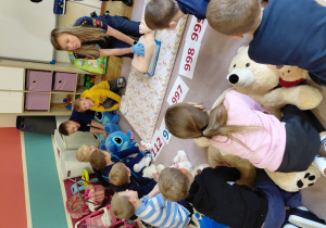 Przedszkolaki siedzą na dywanie wraz z strażniczką miejską, nachylają się nad pluszakami w pozycji do uciśnięć pierwszej pomocy.