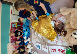 Strażniczka miejska przykrywa dziewczynkę kocem termicznym. Przedszkolaki siedzą na dywanie, przed nimi leżą pluszaki.