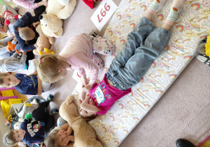 Dziewczynka leży na dywanie z ręką na twarzy, druga dziewczynka siedzi obok niej. Przedszkolaki siedzą na dywanie z pluszakami.