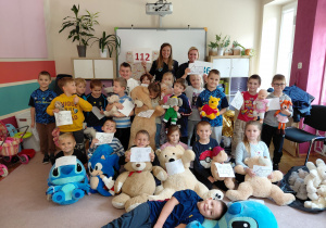 Przedszkolaki z grupy drugiej z pluszakami wraz ze strażniczkami miejskimi siedzą i stoją na dywanie trzymając kolorowanki pojazdów uprzywilejowanych.