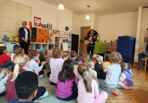 Przedszkolaki siedzą tyłem na dywanie. Kobieta stoi przy szafkach. Mężczyzna z gitarą i sombrero stoi przed dziećmi.