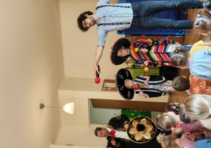 Przedszkolaki siedzą na dywanie. Na środku stoi dwoje dzieci w sombrero i ponczo z instrumentami. Mężczyzna z gitarą i sombrero stoi przed dziećmi. Mężczyzna z wyciągniętym do dzieci mikrofonem stoi obok dwójki dzieci.