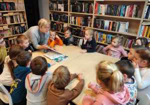 Przedszkolaki siedzą przy stole. Bibliotekarka pokazuje im książki.