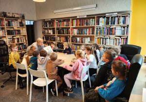 Przedszkolaki siedzą przy stole i na fotelach. Bibliotekarka pokazuje im książki.