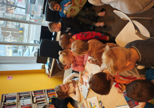 Przedszkolaki siedzą przy stole i na fotelach i oglądają różne książki literatury dziecięcej.