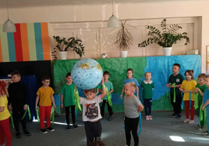 Przedszkolaki stoją na dywanie w półkolu. Na środku stoi dwóch chlopców. Jeden z nich trzyma piłkę - kulę ziemską. Rodzice dzieci siedzą tyłem.