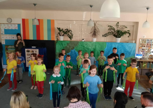 Przedszkolaki z zielonymi i niebieskimi szarfami stoją na dywanie w rozsypce. Rodzice dzieci siedzą tyłem. Z tyłu stoi kobieta.
