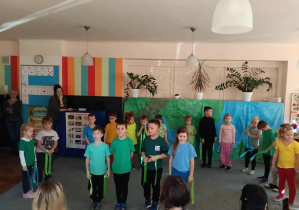 Przedszkolaki stoją na dywanie w półkolu. Na środku stoi czworo dzieci. Rodzice dzieci siedzą tyłem. Za dziećmi stoją dwie kobiety.