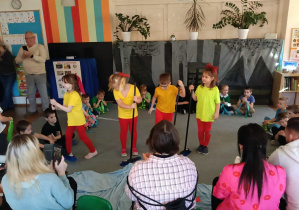 Przedszkolaki siedzą na dywanie w półkolu. Na środku stoi czworo dzieci w żółtych koszulkach ze szczotkami. Rodzice dzieci siedzą tyłem. Za dziećmi stoi mężczyzna i kobieta.