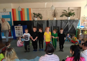 Przedszkolaki siedzą na dywanie w półkolu. Na środku stoi sześcioro dzieci. Rodzice dzieci siedzą tyłem. Za dziećmi stoi mężczyzna.