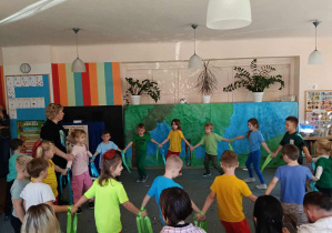 Przedszkolaki idą w kole. obok nich stoi nauczycielka. Rodzice siedzą tyłem.