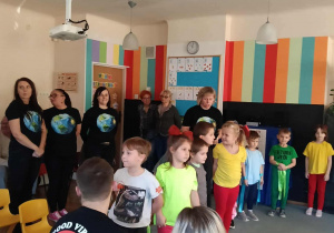 Przedszkolaki stoją na dywanie. Za nimi stoją nauczycielki w koszulkach z Ziemią. Rodzice siedzą tyłem.