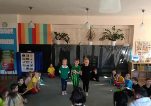 Przedszkolaki siedzą na dywanie w półkolu. Na środku stoi troje dzieci. Rodzice dzieci siedzą tyłem.