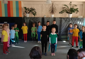 Przedszkolaki stoją na dywanie w półkolu. Na środku stoi dwoje dzieci. Rodzice dzieci siedzą tyłem.