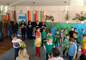 Przedszkolaki stoją na dywanie w rozsypce. Za nimi stoją nauczycielki w koszulkach z kulą ziemską. Rodzice siedzą tyłem.