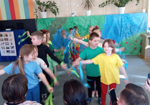 Przedszkolaki stoją w dwóch rzędach na dywanie z wyciągniętymi na boki ramionami. Rodzice siedzą tyłem.