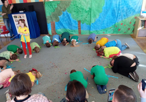 Przedszkolaki siedzą skulone w kole na dywanie. Dziewczynka stoi w środku koła. Kobieta stoi z tyłu za dziećmi.