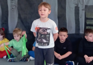 Przedszkolaki siedzą w półkolu na dywanie. Chłopiec stoi na środku.