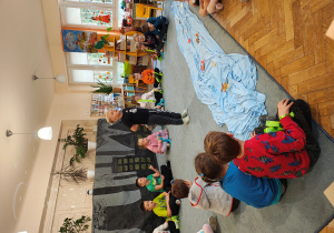 Przedszkolaki siedzą na dywanie w półkolu. Na środku stoi chłopiec. Na dywanie leży niebieski materiał z papierowymi rybkami.