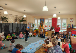 Przedszkolaki siedzą na dywanie w półkolu. Na środku stoi chłopiec. Na dywanie leży niebieski materiał z papierowymi rybkami. Na krzesłach siedzą dzieci. Jedna kobieta stoi przy stołach. Druga kobieta siedzi na krześle.
