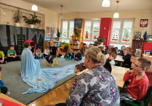 Przedszkolaki siedzą na dywanie w półkolu. Na środku na krzesłach siedzi dwóch chłopców. Na dywanie leży niebieski materiał z papierowymi rybkami. Na krzesłach siedzą dzieci. Jedna kobieta zakłada niebieski materiał na chłopca. Druga kobieta siedzi na krześle.
