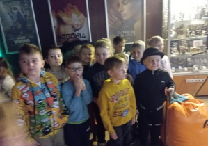 Przedszkolaki stoją na tle plakatów filmowych. Jeden z chłopców ma kapelusz na głowie i laskę w dłoni.