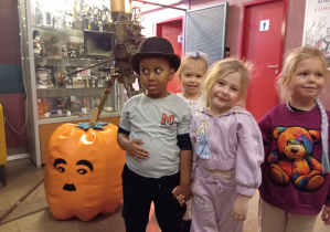 Trzy dziewczynki i jeden chłopiec stoją na tle starej kamery. Chłopiec ma kapelusz na głowie.