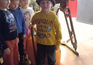 Chłopiec w kapeluszu i z laską stoi na tle kamery. Z boku stoi troje dzieci.