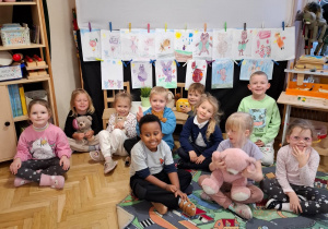 Dzieci siedzą na podłodze i dywanie w ulice, w tle są wywieszone rysunki misiów.