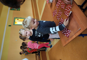 Chłopiec stoi przy stole z wróżbami i odkrywa jeden obrazek. Na stole leżą zakryte obrazki. Za nim stoją dzieci.