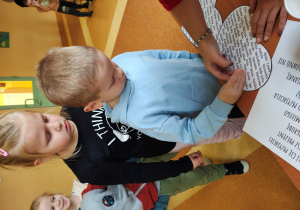 Chłopiec stoi przy stole z wróżbami. Przebija serce z imionami. Za nim stoją dziewczynki.