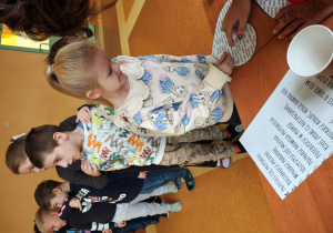 Dziewczynka stoi przy stole z wróżbami. Przebija serce z imionami. Za nią stoją dzieci.