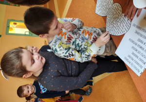 Chłopiec stoi przy stole z wróżbami. Przebija serce z imionami. Za nim stoją dzieci.