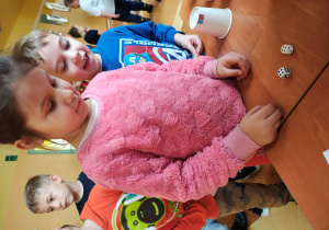 Dziewczynka stoi przy stole z wróżbami. Na stole są dwie kostki do gry. Za nią stoją dzieci.