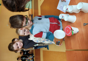 Chłopiec stoi przy stole z wróżbami. Podnosi kubek. Za nim stoją dzieci.