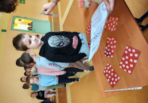 Chłopiec stoi przy stole z wróżbami. Na stole leżą zakryte obrazki. Za nim stoją dzieci.