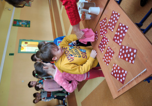 Chłopiec stoi przy stole z wróżbami. Na stole leżą zakryte obrazki. Chłopiec losuję kartkę z torebki. Za nim stoją dzieci.