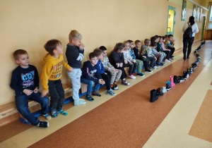 Dzieci siedzą na ławce i stoją na podłodze. Na podłodze w linii ustawione są buty.