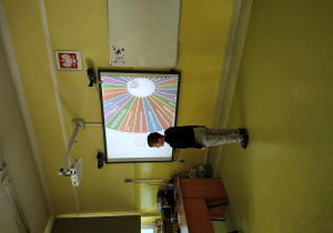 Chłopiec stoi przy tablicy multimedialnej. Na tablicy jest koło z zapisanymi nazwami zawodów.