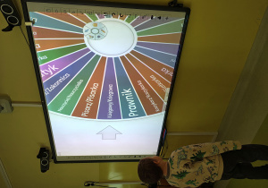 Chłopiec stoi przy tablicy multimedialnej. Na tablicy jest koło z zapisanymi nazwami zawodów.