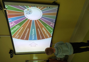 Chłopiec stoi przy tablicy multimedialnej. Na tablicy jest koło z zapisanymi nazwami zawodów.