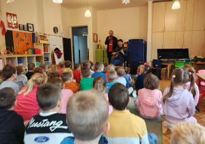 Mężczyzna w czarnej chustce siedzi na krześle z mandoliną. Za nim stoi mężczyzna z mikrofonem. Przed nimi, na dywanie siedzą przedszkolaki.