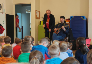 Mężczyzna w czarnej chustce siedzi na krześle z mandoliną. Za nim stoi mężczyzna z mikrofonem. Przed nimi, na dywanie siedzą przedszkolaki.