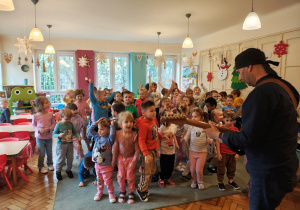 Mężczyzna w czarnej chustce stoi z gitarą elektryczną. Przed nim, na dywanie stoją przedszkolaki.