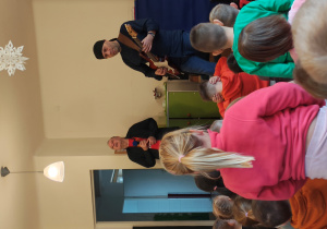 Mężczyzna w czarnej chustce stoi z gitarą elektryczną. Obok niego stoi mężczyzna z mikrofonem. Przed nimi, na dywanie siedzą przedszkolaki.