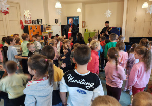 Mężczyzna w czarnej chustce stoi z gitarą elektryczną. Przed nim stoi mężczyzna z mikrofonem. Przed nimi, na dywanie stoją przedszkolaki.