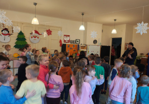 Mężczyzna w czarnej chustce stoi z gitarą elektryczną. Przed nim, na dywanie stoją przedszkolaki.